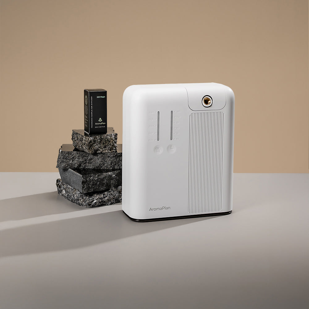 A clean, white aroma diffuser displayed on a neutral, minimalistic grey surface. The diffuser is positioned next to stacked black stones, creating a stylish contrast. A scent cartridge is placed atop the stones, showcasing the AromaPlan logo. The background is a soft beige, enhancing the elegant design of the product.