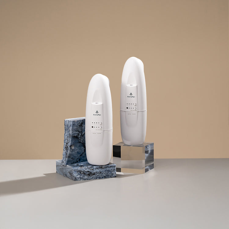 A collection of sleek, modern diffusers with varying sizes and shapes, placed on minimalist stone stands. The diffusers are placed on a light grey surface with a warm beige background.